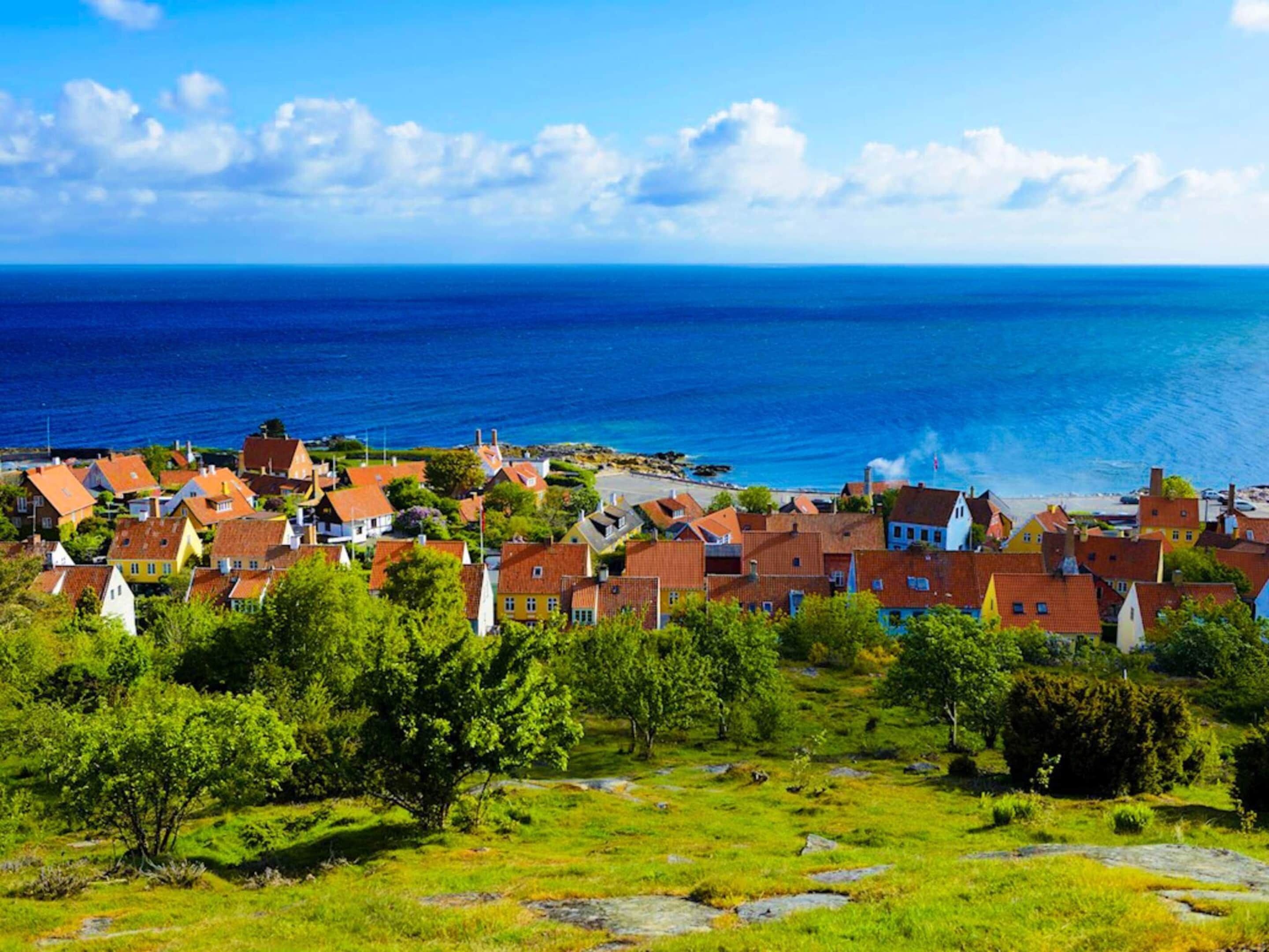 A coastal village with traditional red-roofed houses, a clear view of the sea, and a grassy foreground.