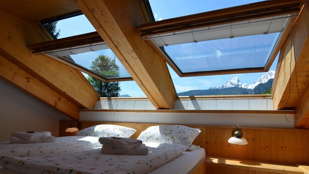 A bedroom with a bed, pillows, and a bedside table. There are skylights allowing natural light and a view of mountains.