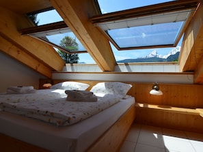 A bedroom with a bed, pillows, and a bedside table. There are skylights allowing natural light and a view of mountains.