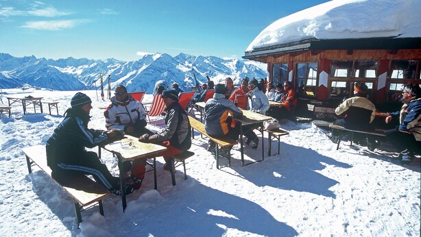 Un groupe de personnes est assis à des tables à l’extérieur, avec des montagnes enneigées en arrière-plan.