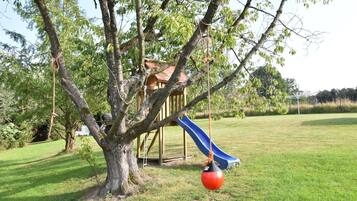 A playground with a slide and swings, surrounded by a grassy field and trees.