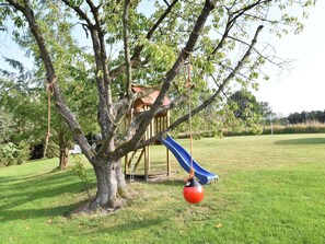 Une aire de jeux avec un toboggan et des balançoires, entourée d’un champ d’herbe et d’arbres.
