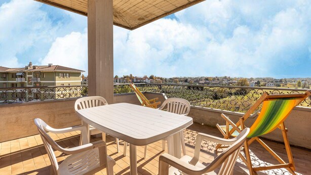 A balcony with a table and chairs, a colorful beach chair, and a view of a residential area. - Nice Apartment in Tuoro sul Trasimeno With Pool (Tuoro sul Trasimeno)