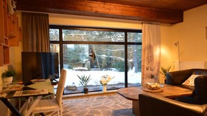 A cozy living room with a stone floor, a television, a glass table, and a large window with a view of trees and snow. - Cosy Apartment in Manderscheid With Balcony (Manderscheid)