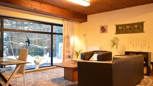 A cozy living room with a stone-patterned floor, a wooden ceiling, a large window with a view of snow-covered trees, a black leather sofa, a glass coffee table, and a small side table with a plant. - Cosy Apartment in Manderscheid With Balcony (Manderscheid)
