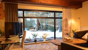 A cozy living room with a stone-patterned floor, a large window with a view of a snowy landscape, a television, and a dining area with a table and chairs. - Cosy Apartment in Manderscheid With Balcony (Manderscheid)