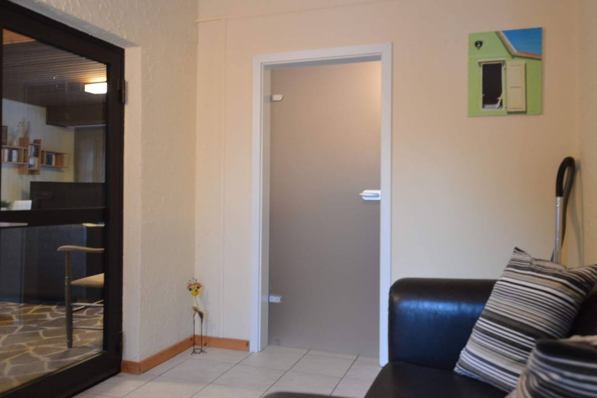 A living room with a black leather sofa, a glass door leading to a kitchen, and a green wall with a small window.
