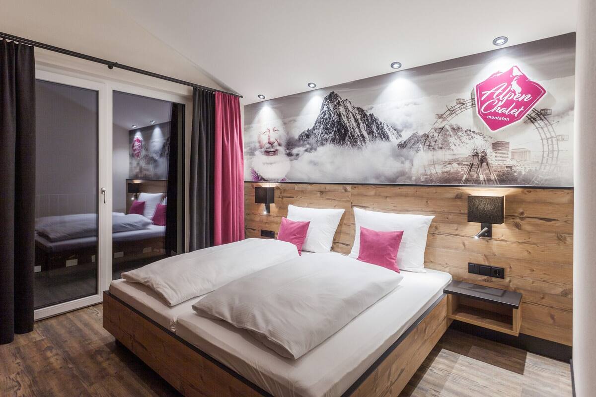 A hotel room with a bed, a wooden headboard, a wall-mounted lamp, and a large window with curtains.