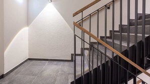 A staircase with a wooden handrail and black metal balusters, leading to a landing with a textured white wall and a recessed light fixture. - Chalet Montafon in Gaschurn With Sauna (Gaschurn)