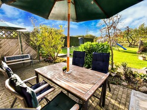 Eine Terrasse im Freien mit einem Holztisch und -stühlen, einem grünen Sonnenschirm und einem Spielbereich im Hintergrund.