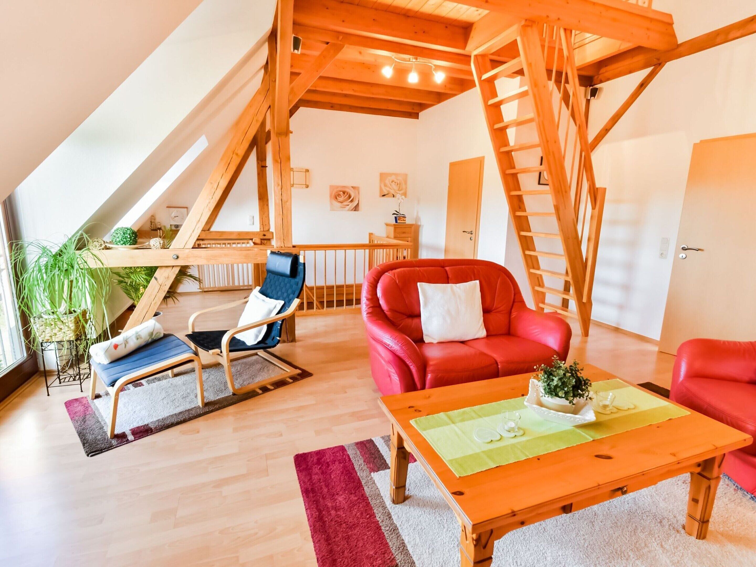A spacious loft with a high ceiling, wooden beams, a red leather sofa, a blue chair, and a wooden table.