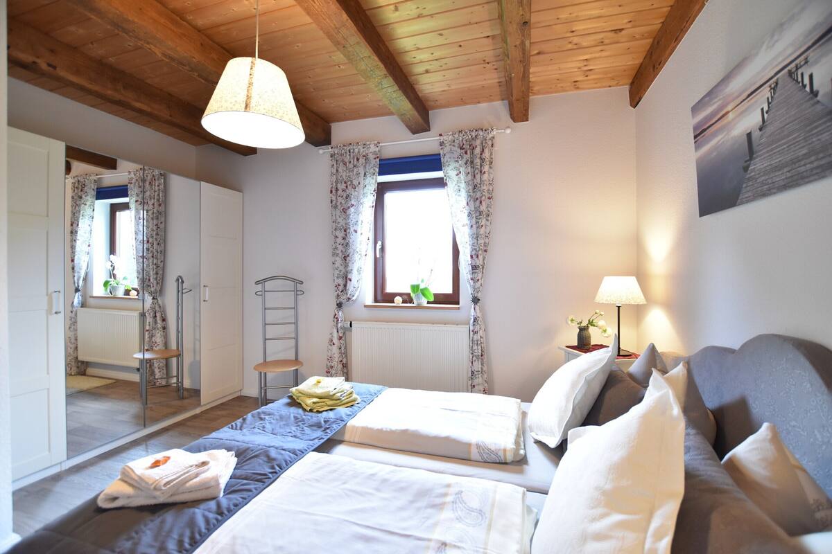 A well-lit bedroom with two beds, a window with curtains, a bedside table with a lamp, and a wooden ceiling.