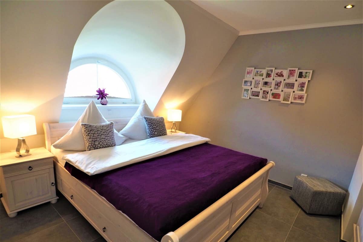 A bedroom with a bed, two bedside tables, a window with a view, and a wall with pictures.