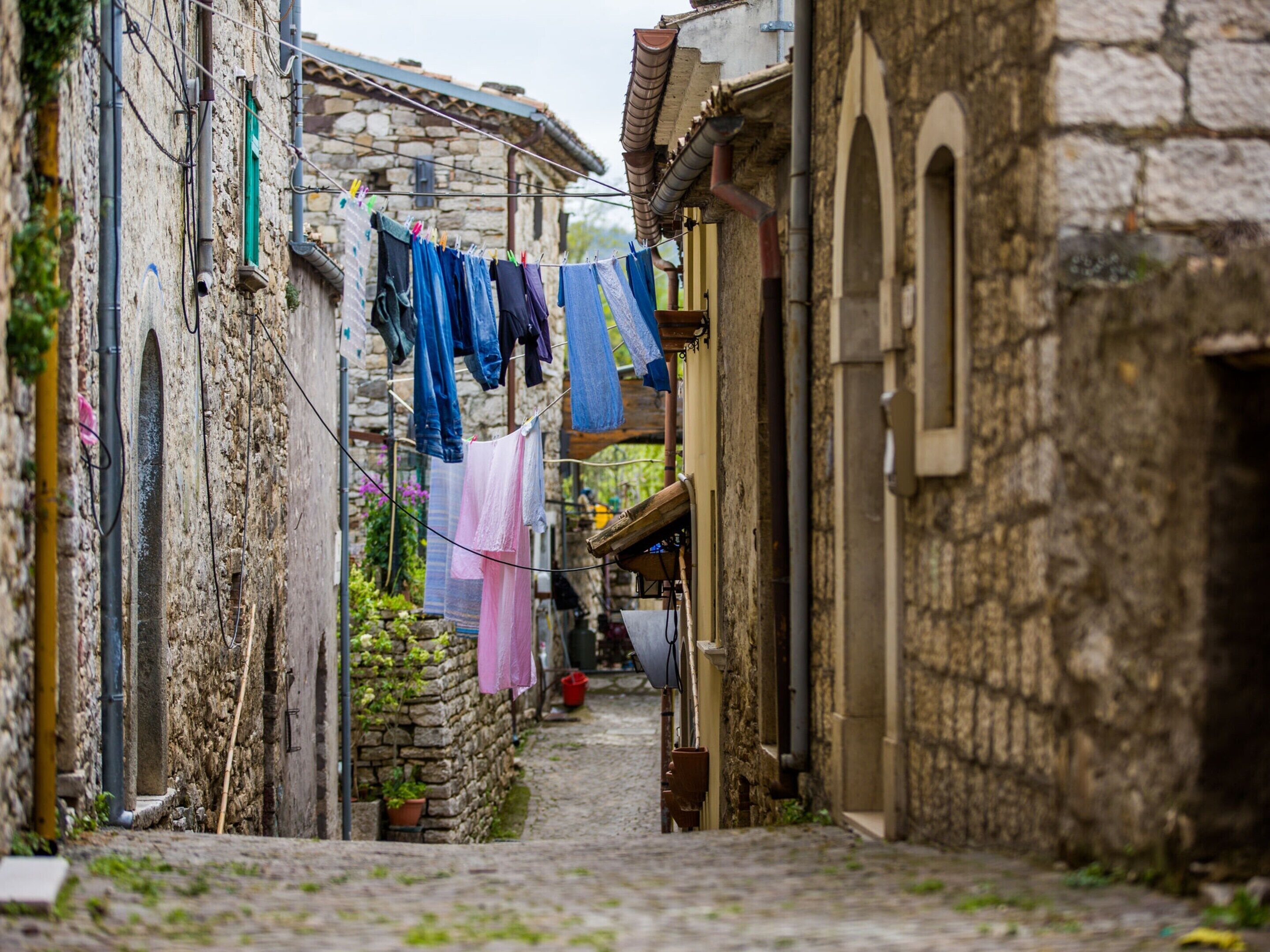 A narrow cobblestone street with stone buildings, laundry hanging on a line, and a small garden area.