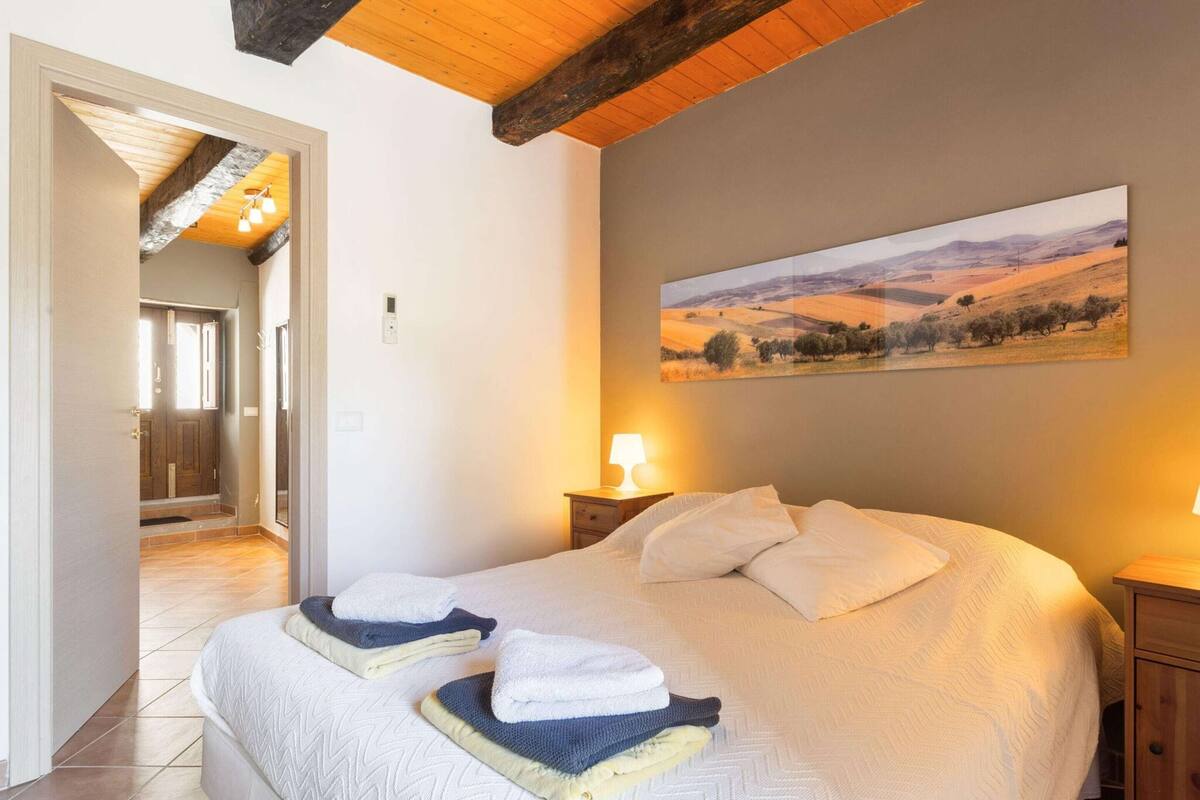 A bedroom with a large bed, a painting of a landscape, and a wooden ceiling.