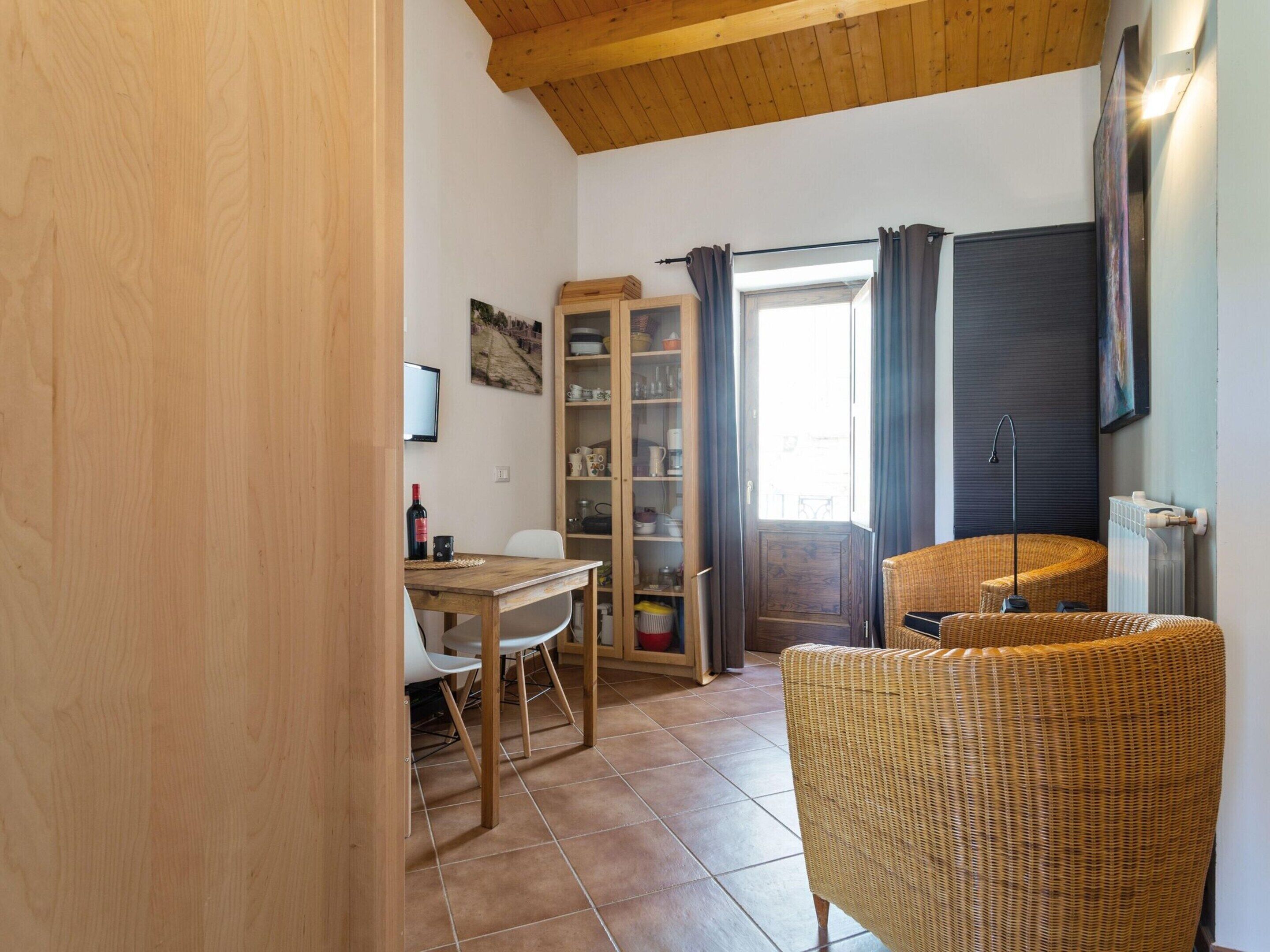 A cozy room with a wooden ceiling, a dining table with chairs, a wicker chair, a bookshelf, and a radiator.