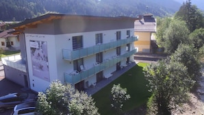 En bygning med flere etager, med balkon på anden sal, omgivet af grøn vegetation og bjerge i baggrunden.