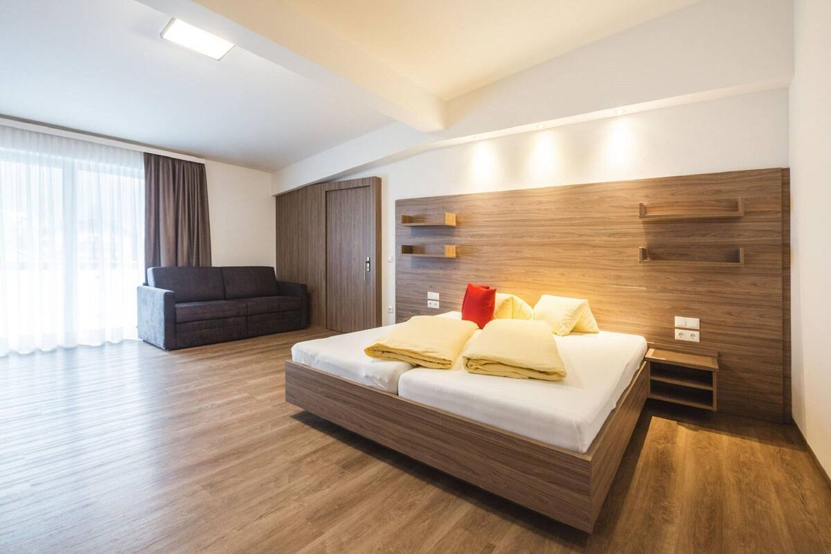 A modern hotel room with a wooden bed, a sofa, and a large window with curtains.