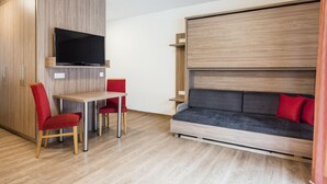 En kompakt opholdsstue med et fjernsyn monteret på en trævæg, et lille spisebord med røde stole og en sofa med røde puder.