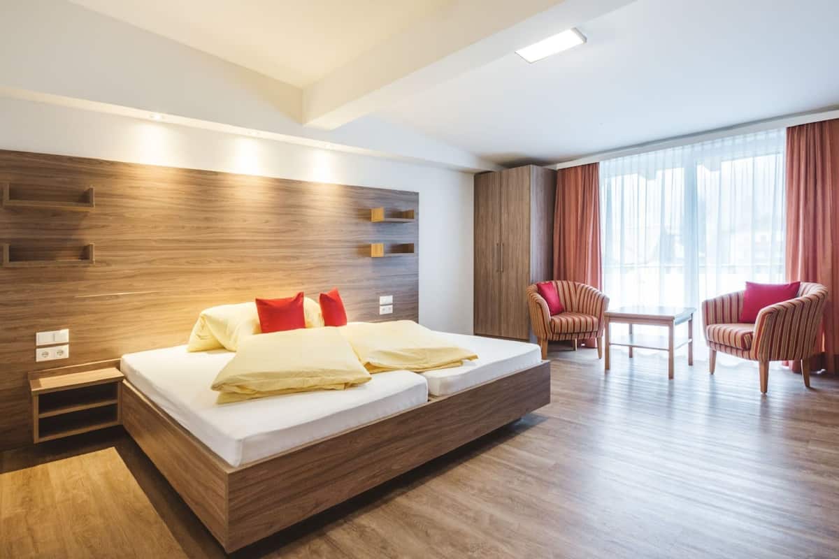 A modern hotel room with a large bed, a wooden headboard, a seating area with two chairs, and a large window with curtains.