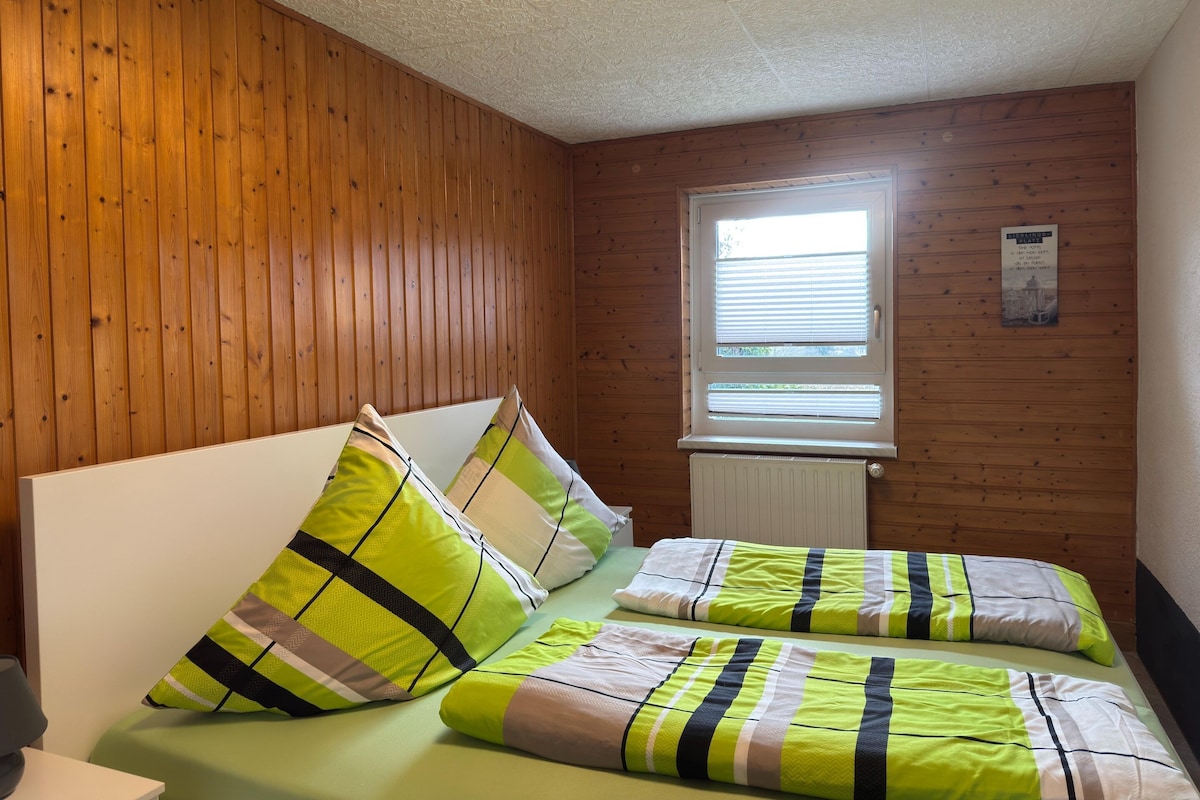 A room with wooden panel walls, a window with blinds, a bed with striped bedding, and a radiator.