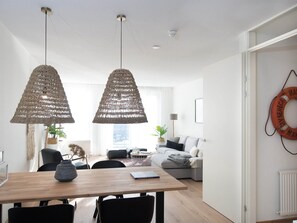 A modern living room with a wooden dining table, two hanging woven light fixtures, a grey sofa, and a white wall with a life preserver decoration. - Modern Apartment Close to the Beach and the sea (The Hague)