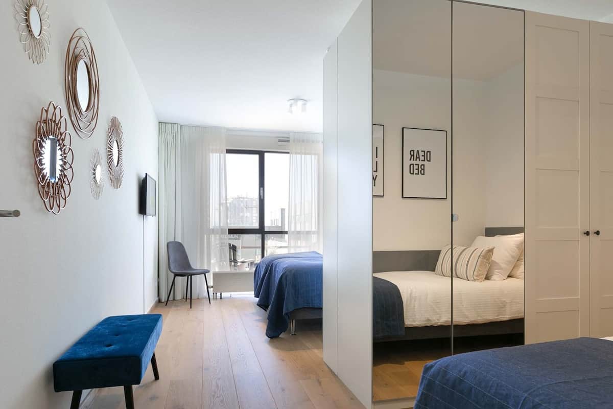 A modern bedroom with a large bed, a smaller bed, a bench, and a chair. There are decorative mirrors on the wall and a window with curtains.