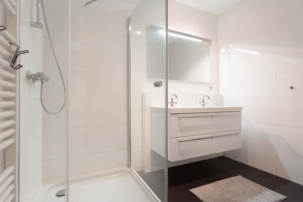 Un baño moderno con ducha de vidrio, tocador blanco con espejo y piso oscuro.