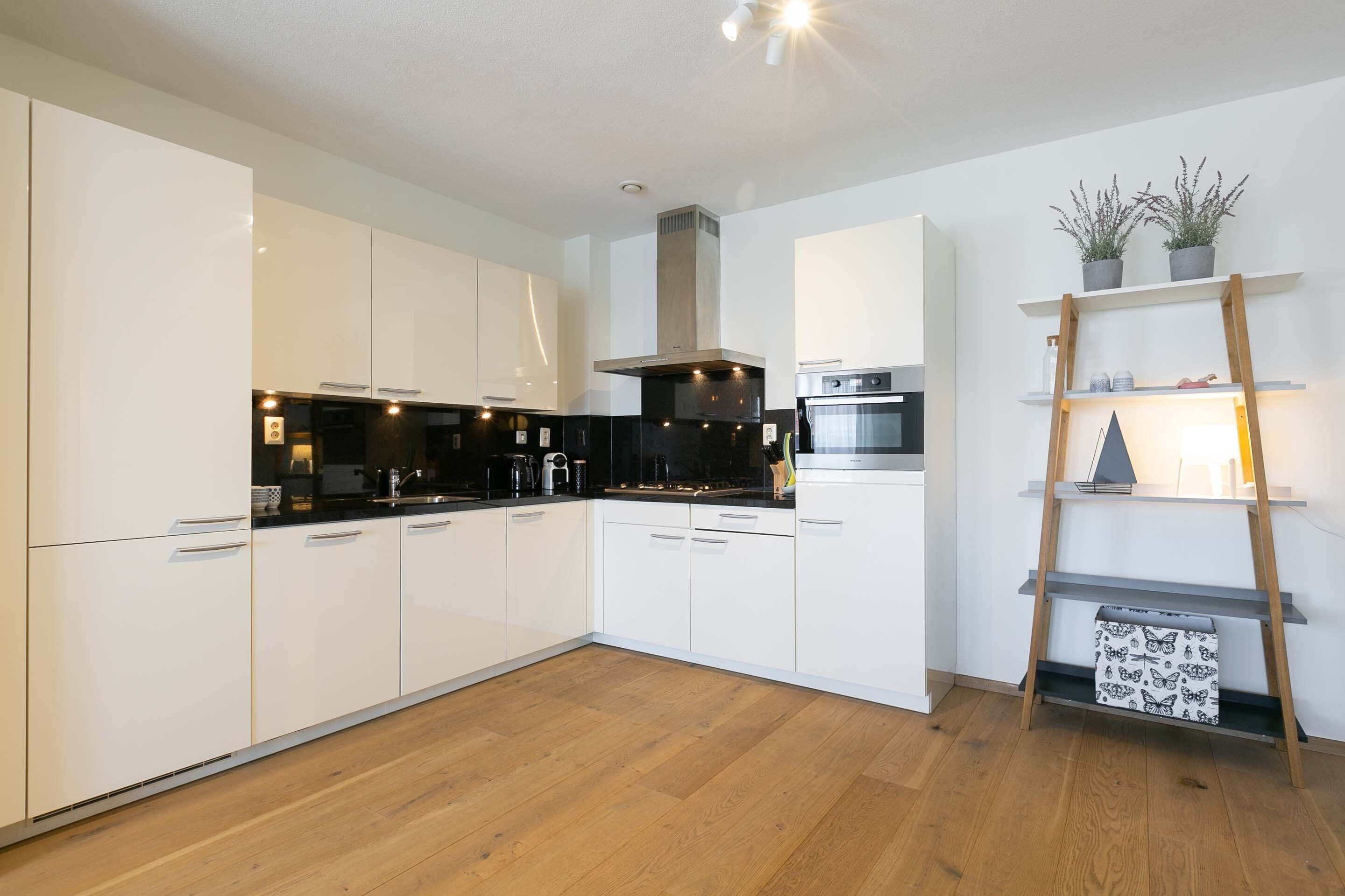 Una cocina moderna con armarios blancos, encimera negra y suelo de madera. Hay un horno de acero inoxidable y un microondas en la encimera. Una estantería de madera con plantas está apoyada en la pared.