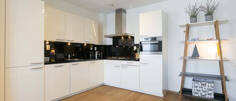 Una cocina moderna con armarios blancos, encimera negra y suelo de madera. Hay un horno de acero inoxidable y un microondas en la encimera. Una estantería de madera con plantas está apoyada en la pared.