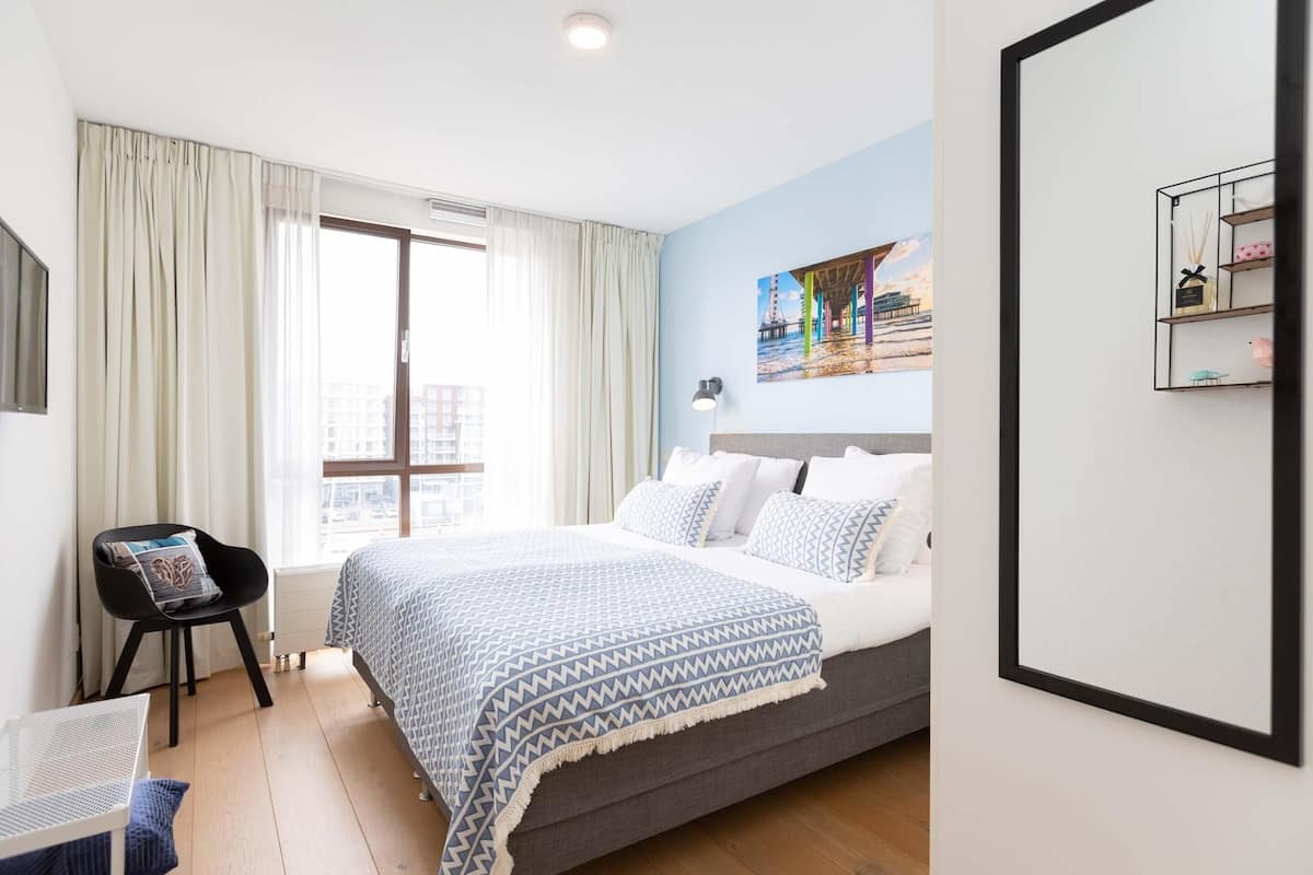 A bedroom with a bed, a chair, a window with curtains, a painting on the wall, and a shelf with items.