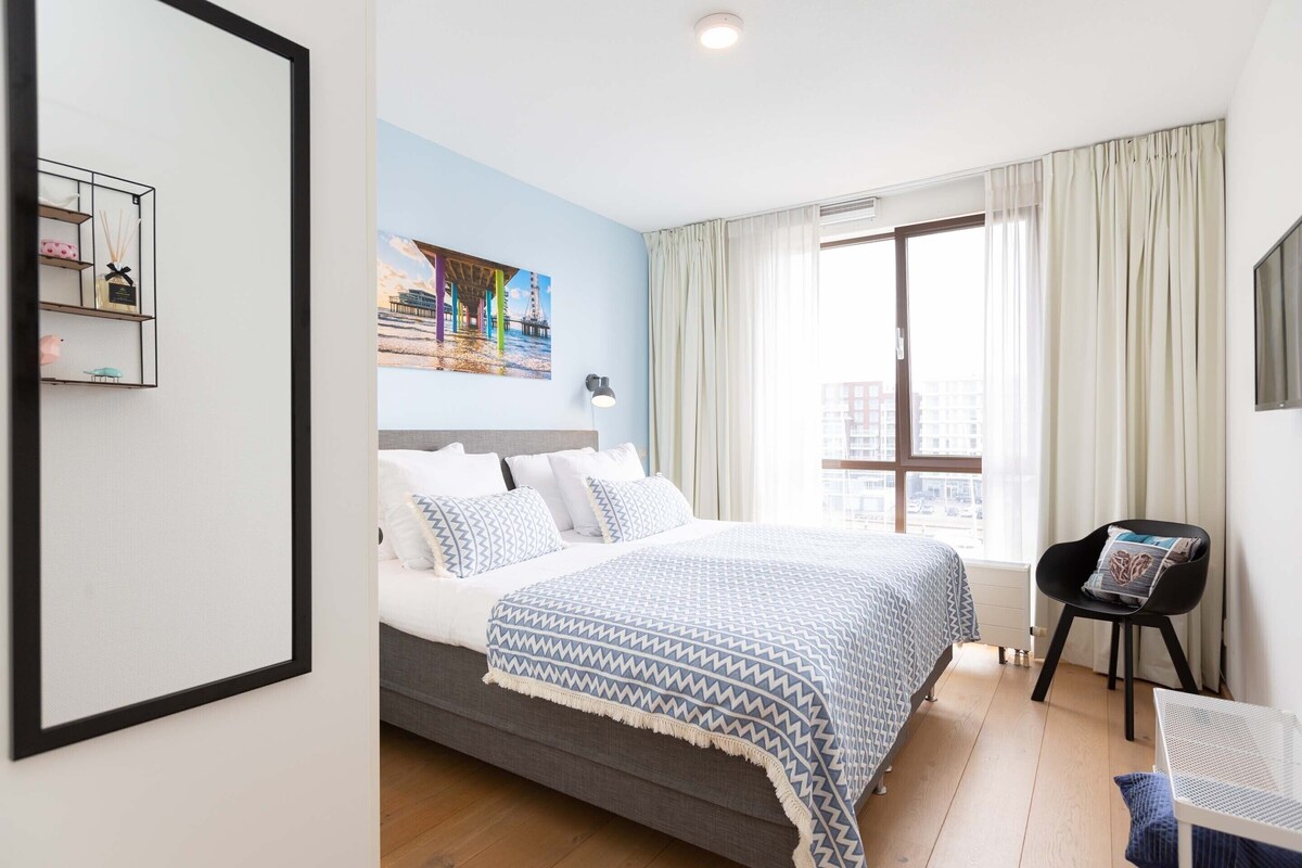A bedroom with a bed, a chair, a window with curtains, and a painting on the wall.