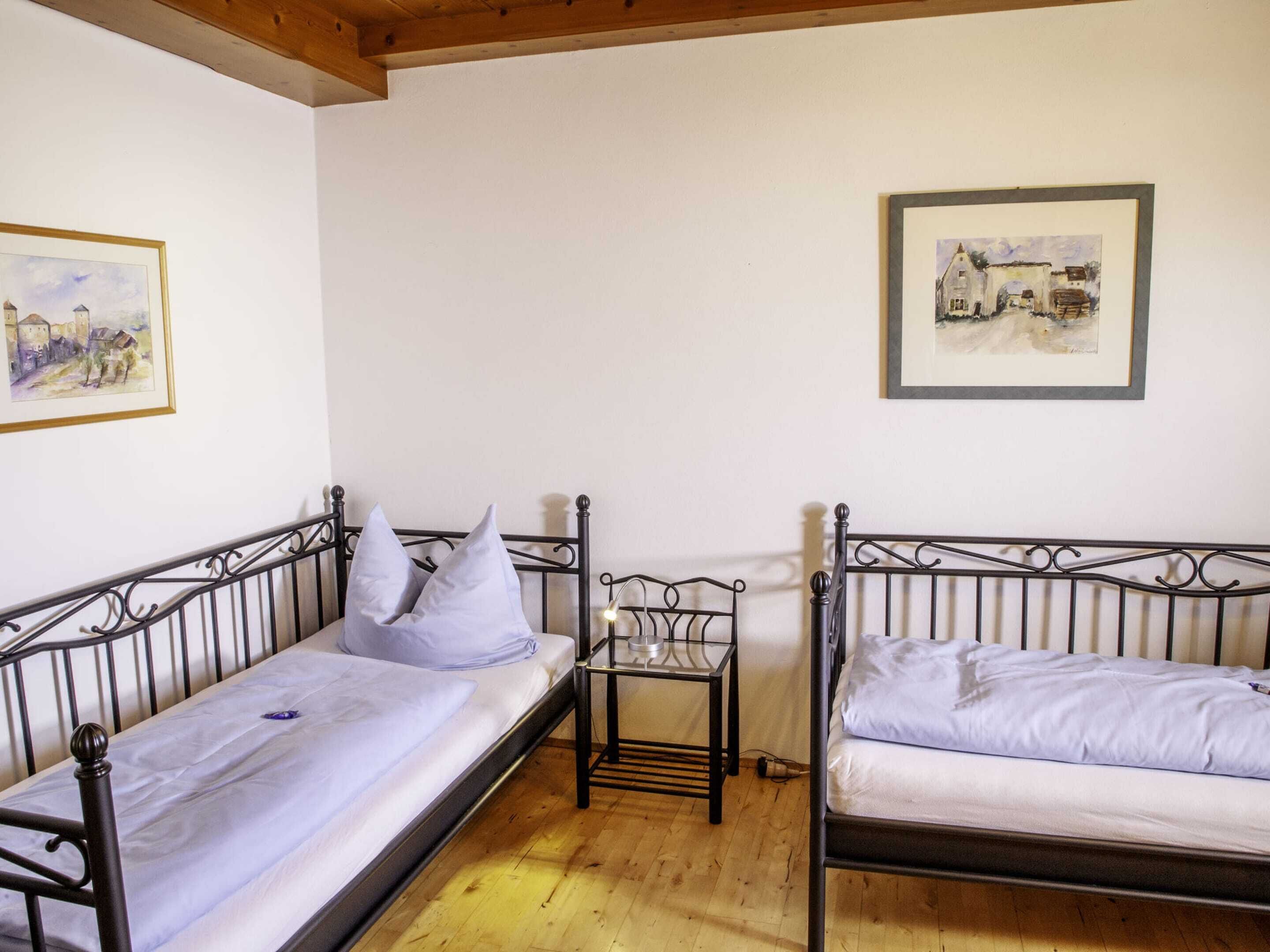 Een kamer met twee metalen bedden, witte beddengoed, een tafeltje met een lamp en twee ingelijste foto's aan de wand.