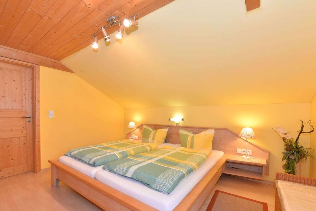 A cozy bedroom with a wooden ceiling, a large bed with a striped bedspread, two bedside tables with lamps, and a small plant on the right.