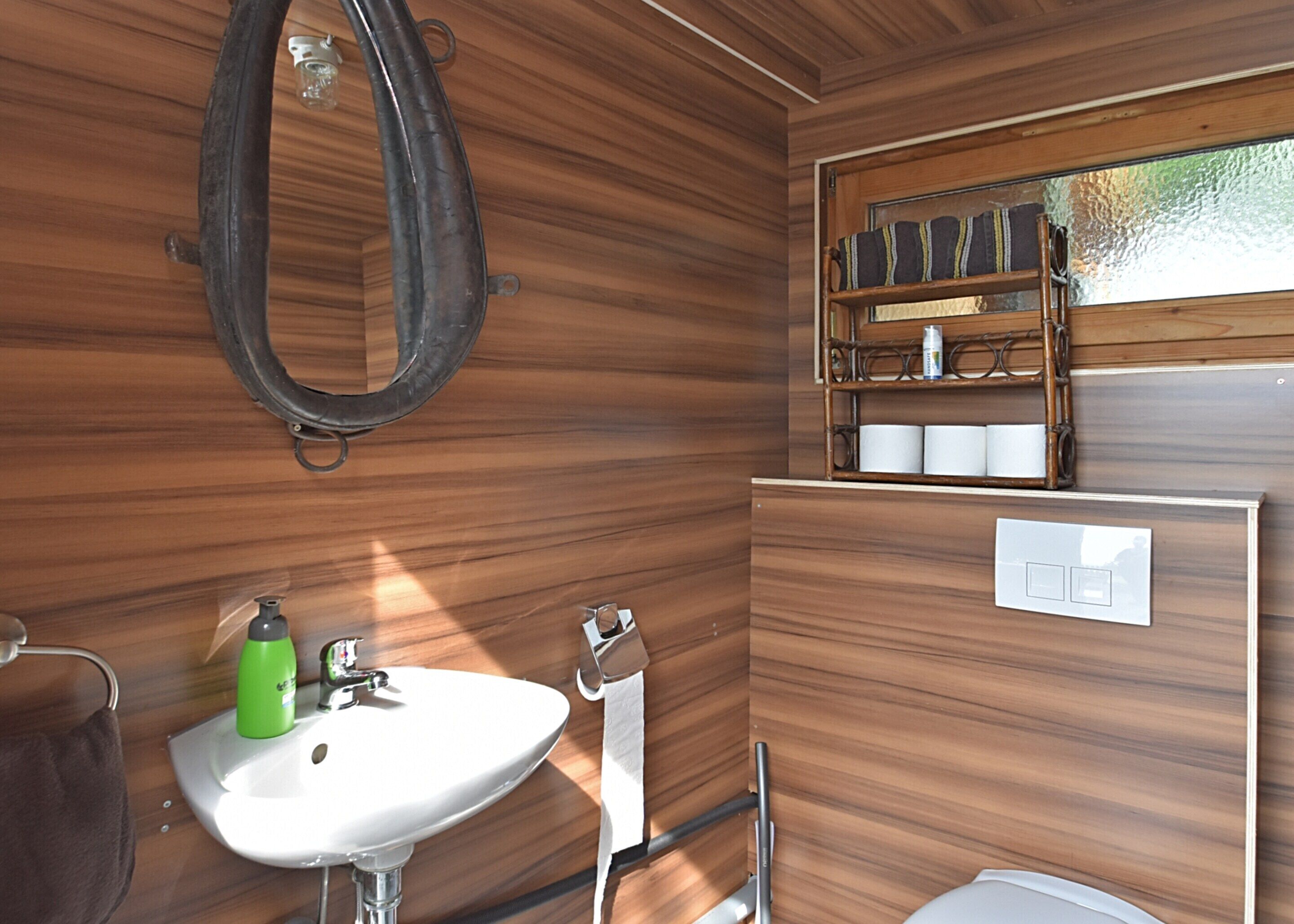 Un baño pequeño con pared de madera, un lavamanos blanco, una botella verde, un perchero, un inodoro y un estante con artículos de aseo.