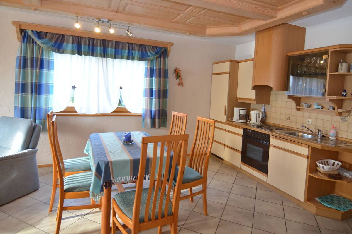 A compact kitchen with wooden cabinets, a dining table with chairs, and a window with curtains.