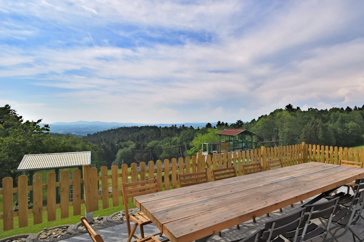 A wooden picnic table with chairs, a wooden fence, and a gazebo in a park with a view of a forest and distant mountains.