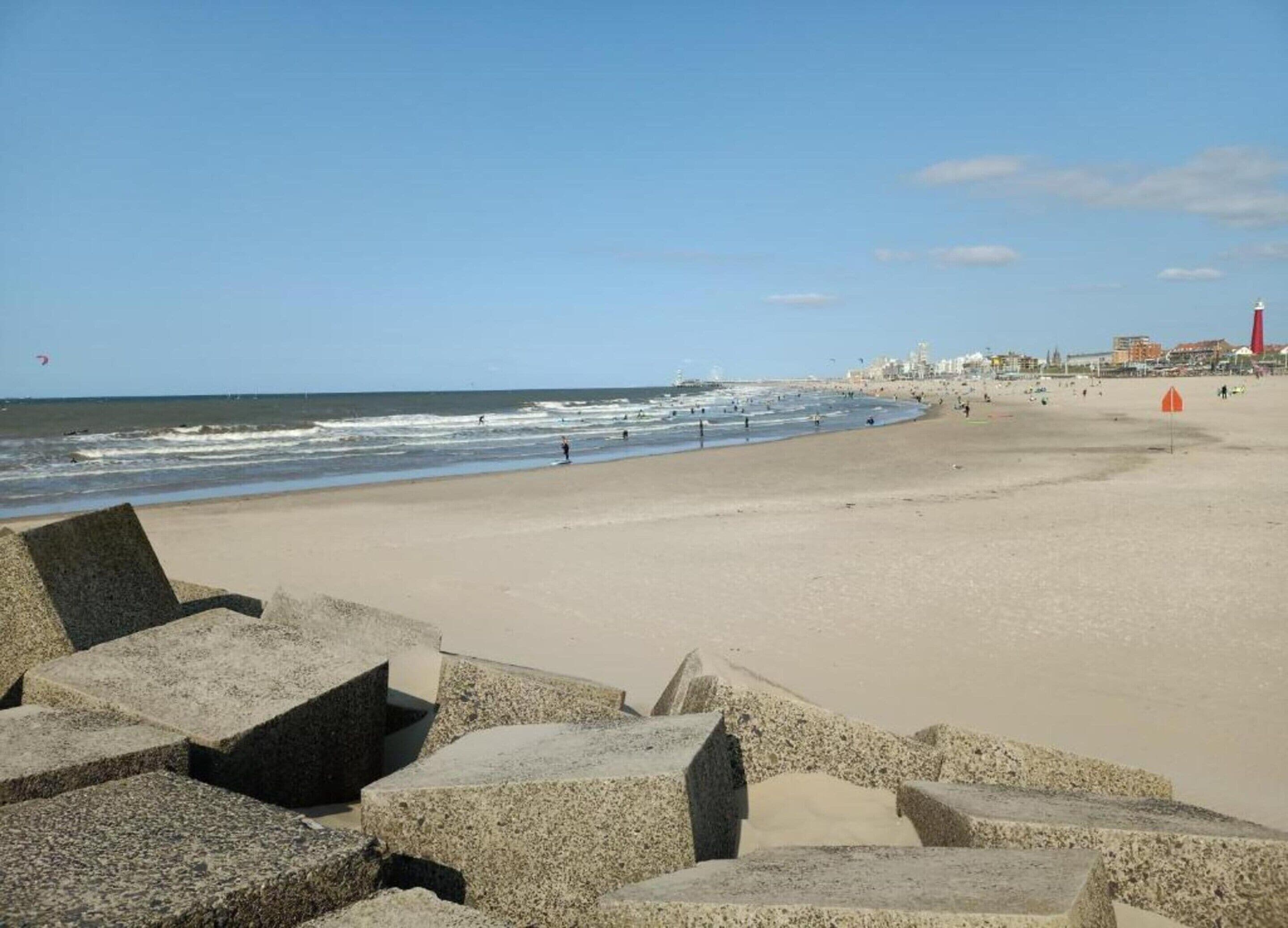 A wide sandy beach with waves, a lighthouse, and a cityscape in the background.