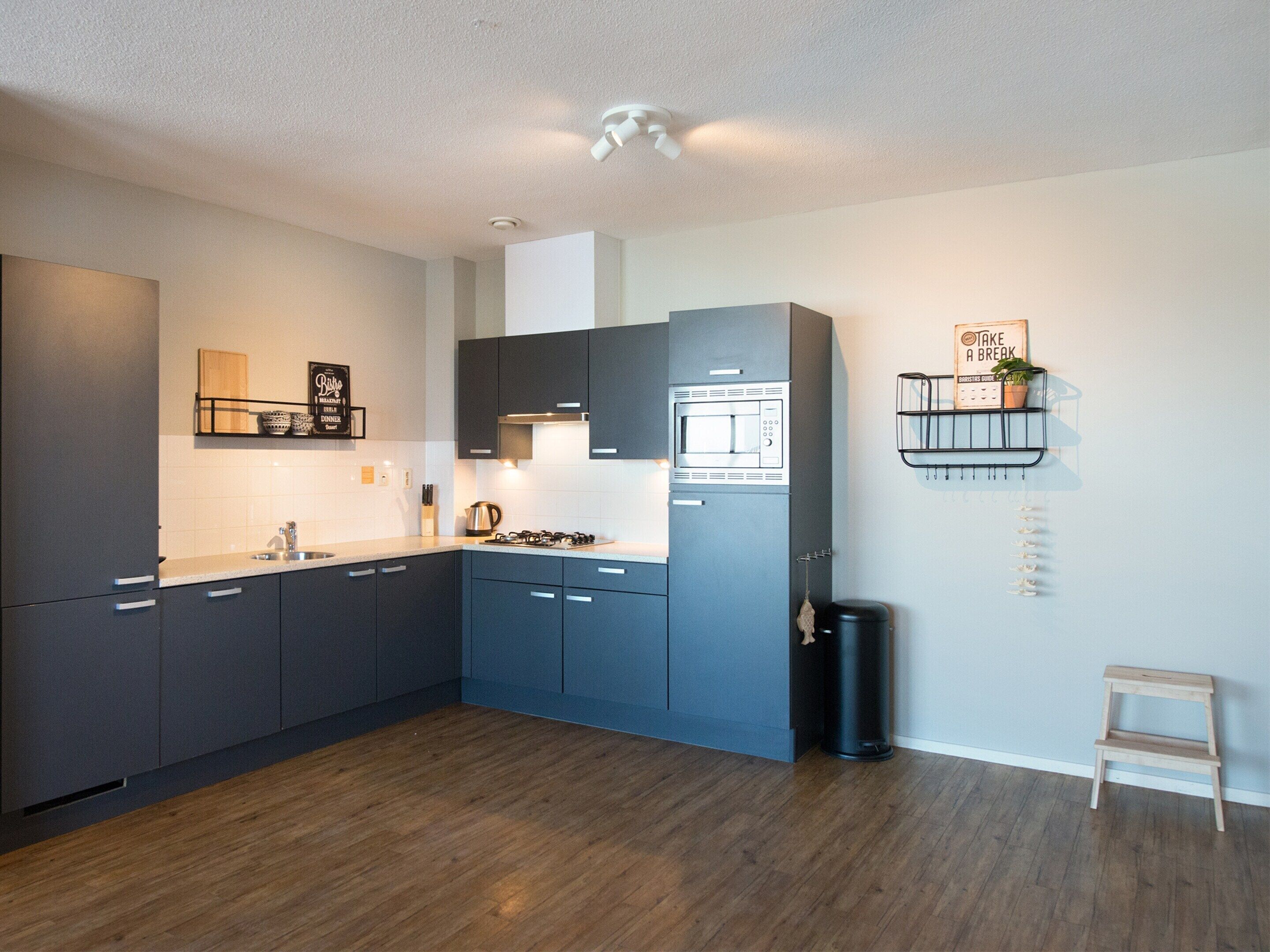 A modern kitchen with dark cabinets, a stainless steel microwave, and a gas stove. There is a small trash bin and a wooden stool near the wall.