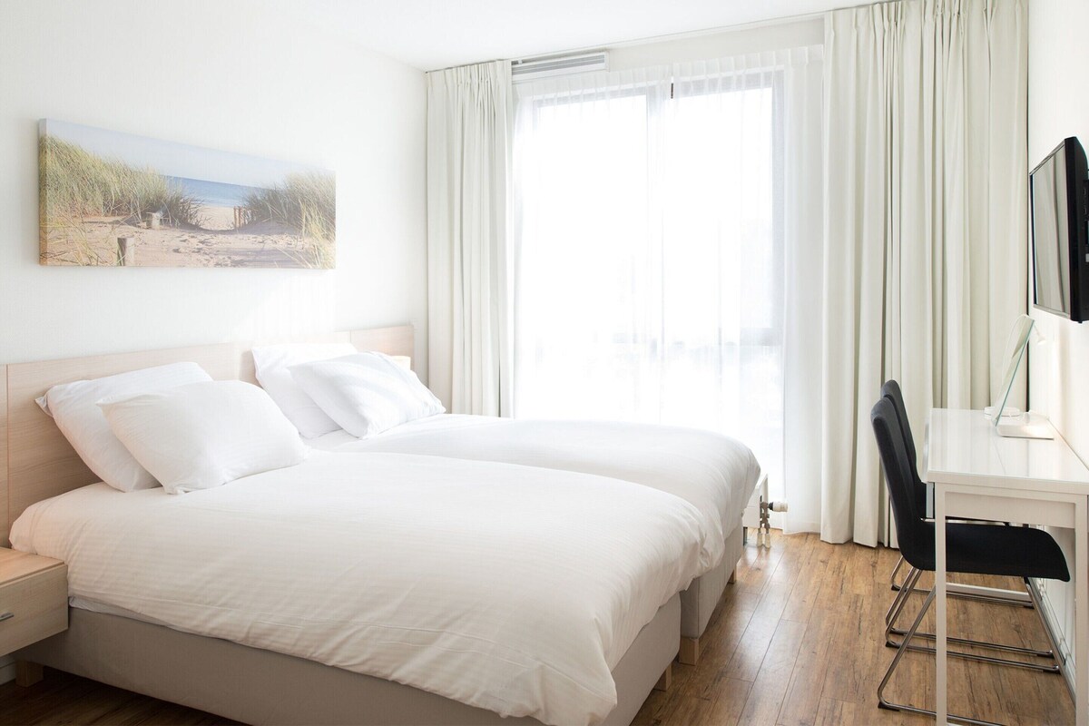 A hotel room with a bed, a desk with a chair, a TV, and a painting on the wall.