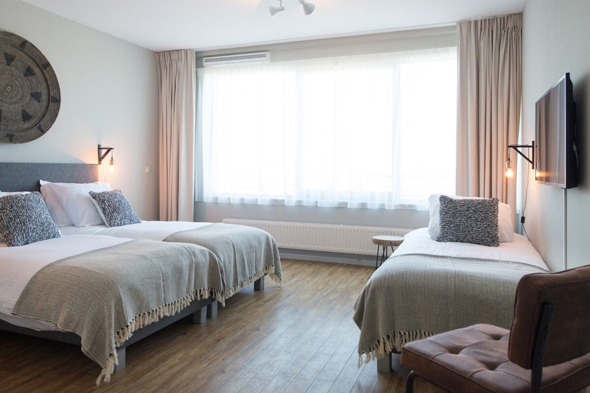 A hotel room with two beds, a wooden floor, a large window with curtains, a round wall decoration, and a mounted TV.