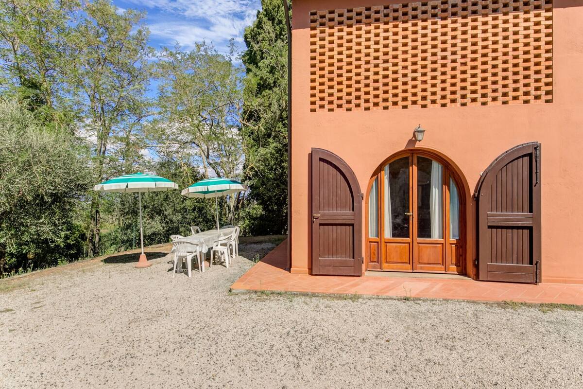 A building with a gravel driveway, two wooden doors, and a window with curtains. There are two white chairs and two umbrellas in front.