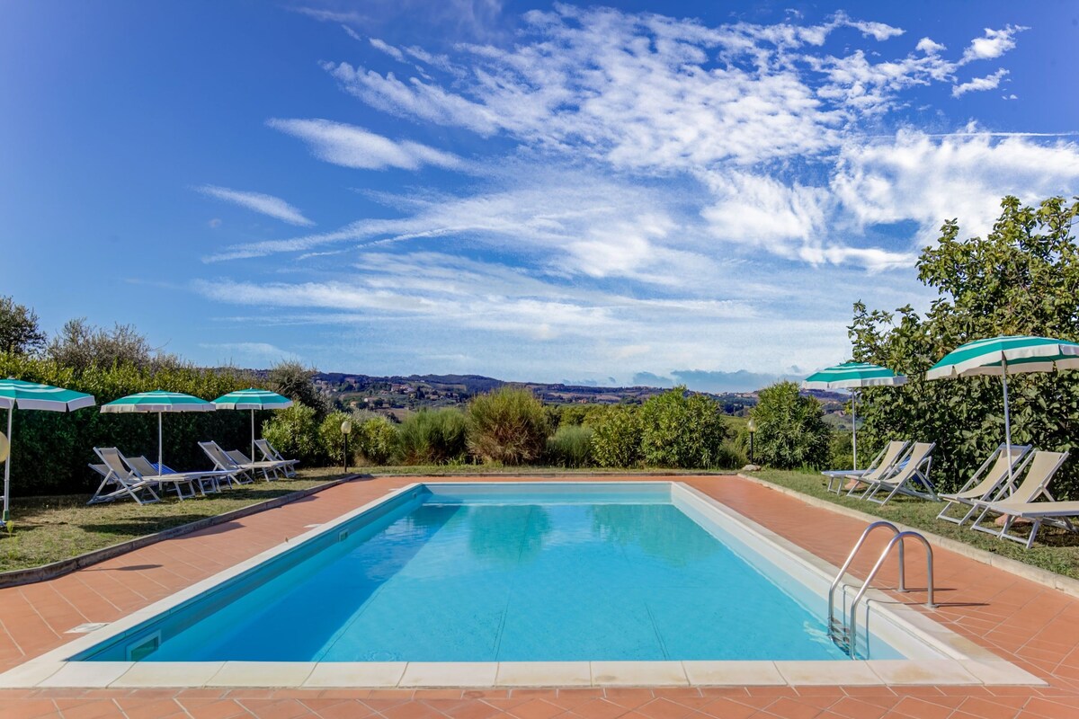 A rectangular swimming pool with a tiled border, surrounded by lounge chairs and umbrellas, with a view of distant hills under a partly cloudy sky.