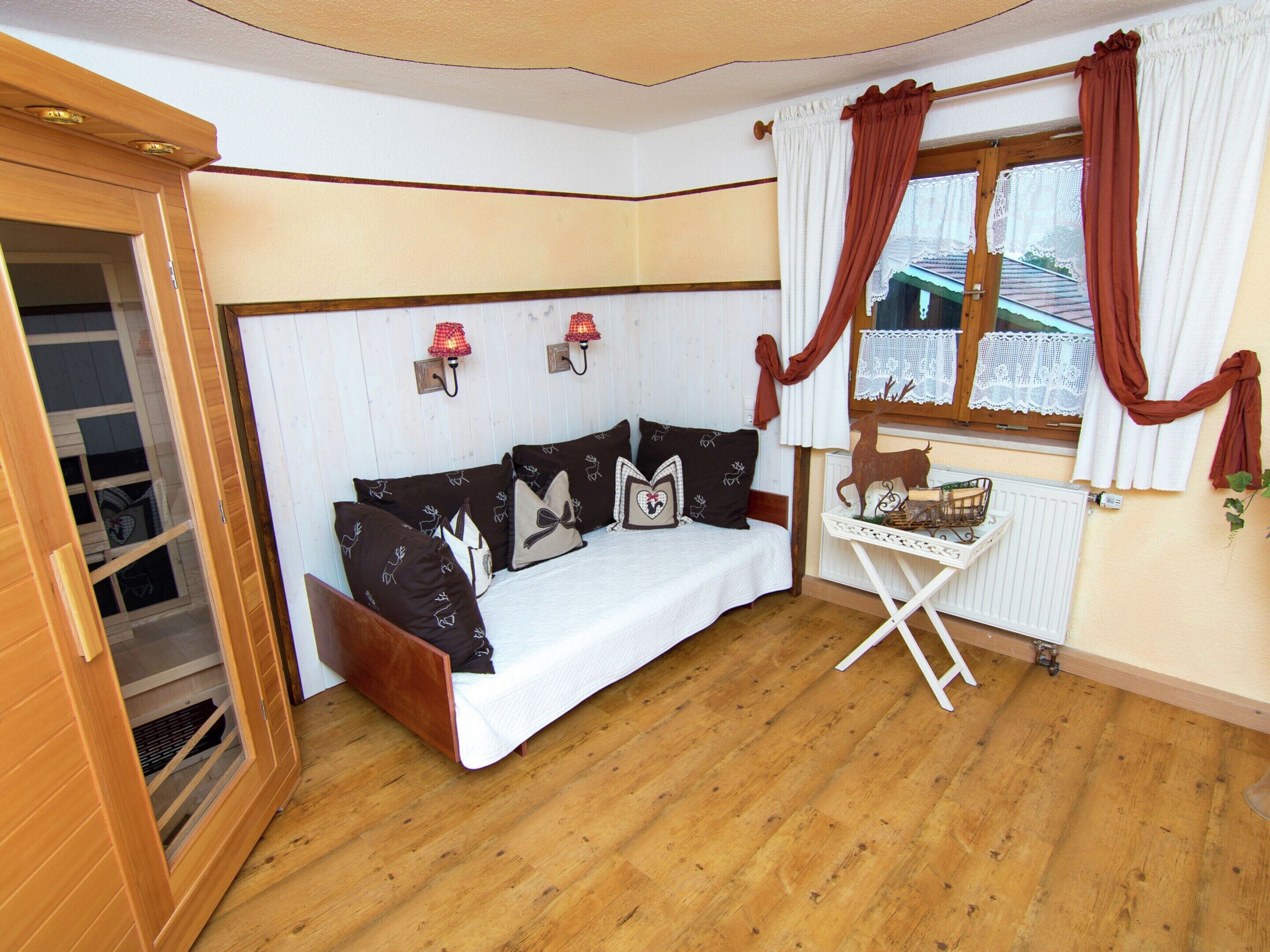 A cozy room with a wooden floor, a white sofa with patterned cushions, a small table with a basket, and a window with curtains.