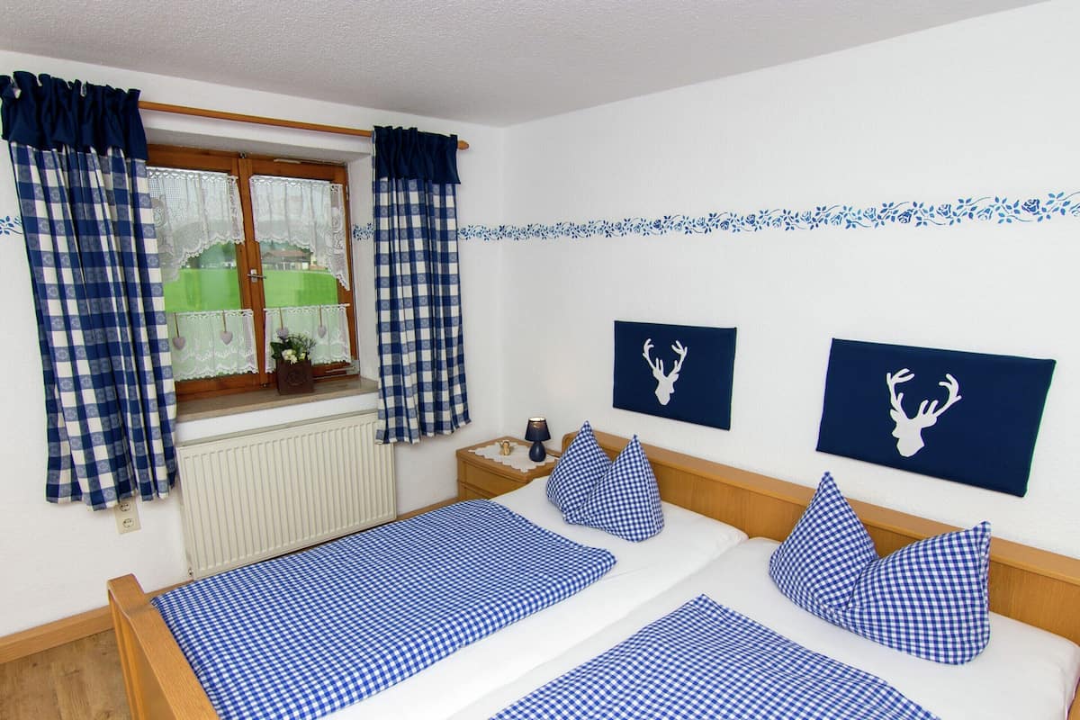 A bedroom with two beds, checkered bedding, a window with curtains, a radiator, a bedside table with a lamp, and two wall-mounted deer head decorations.