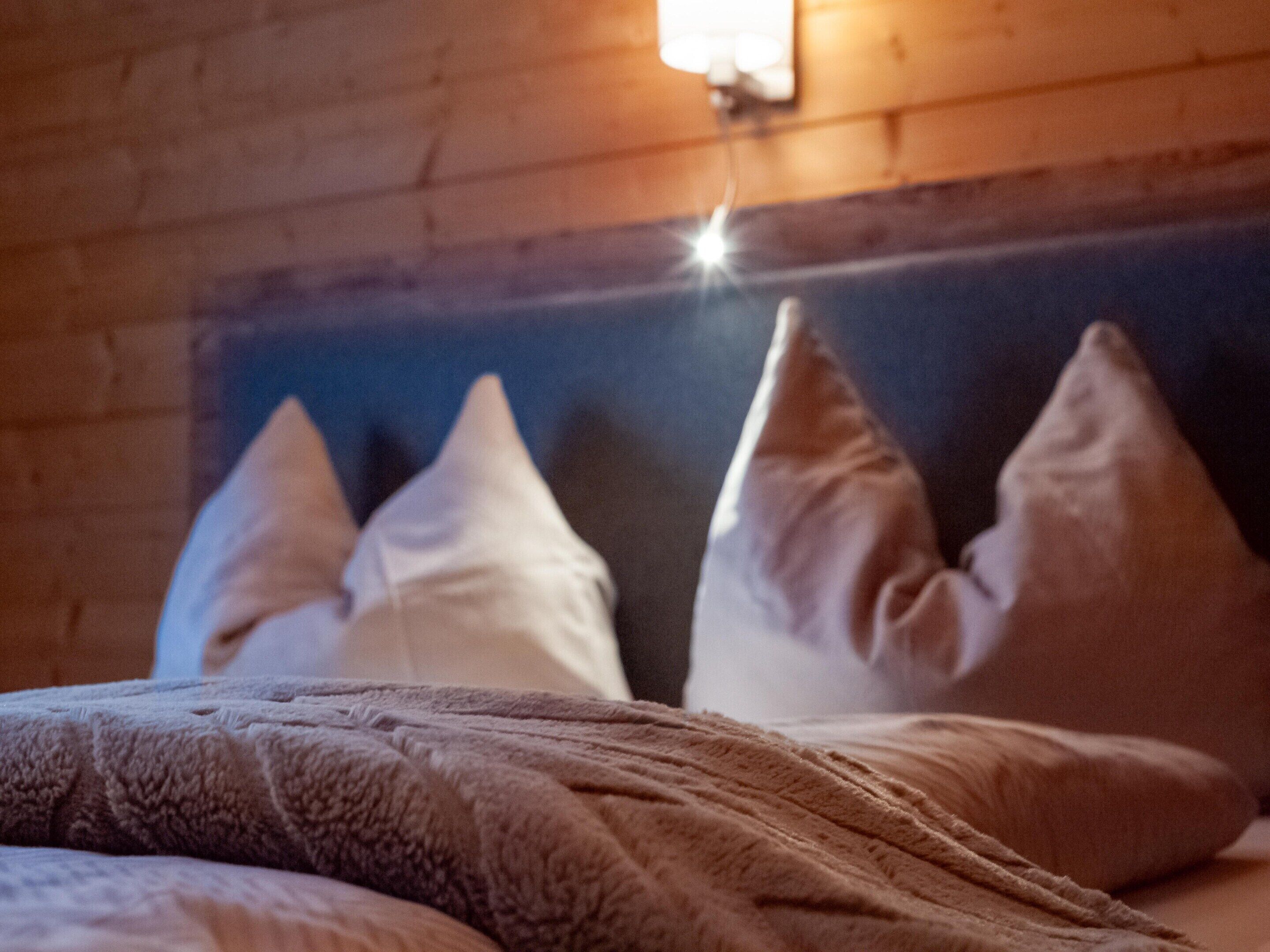 A bed with a textured blanket and three pillows against a wooden wall with a light fixture.