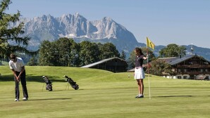 A golf course with a mountainous backdrop, a flag on a flagstick, and a building with a sloped roof. - Modern Apartment in Kitzbuhel Near Ski Area (Kitzbuhel)