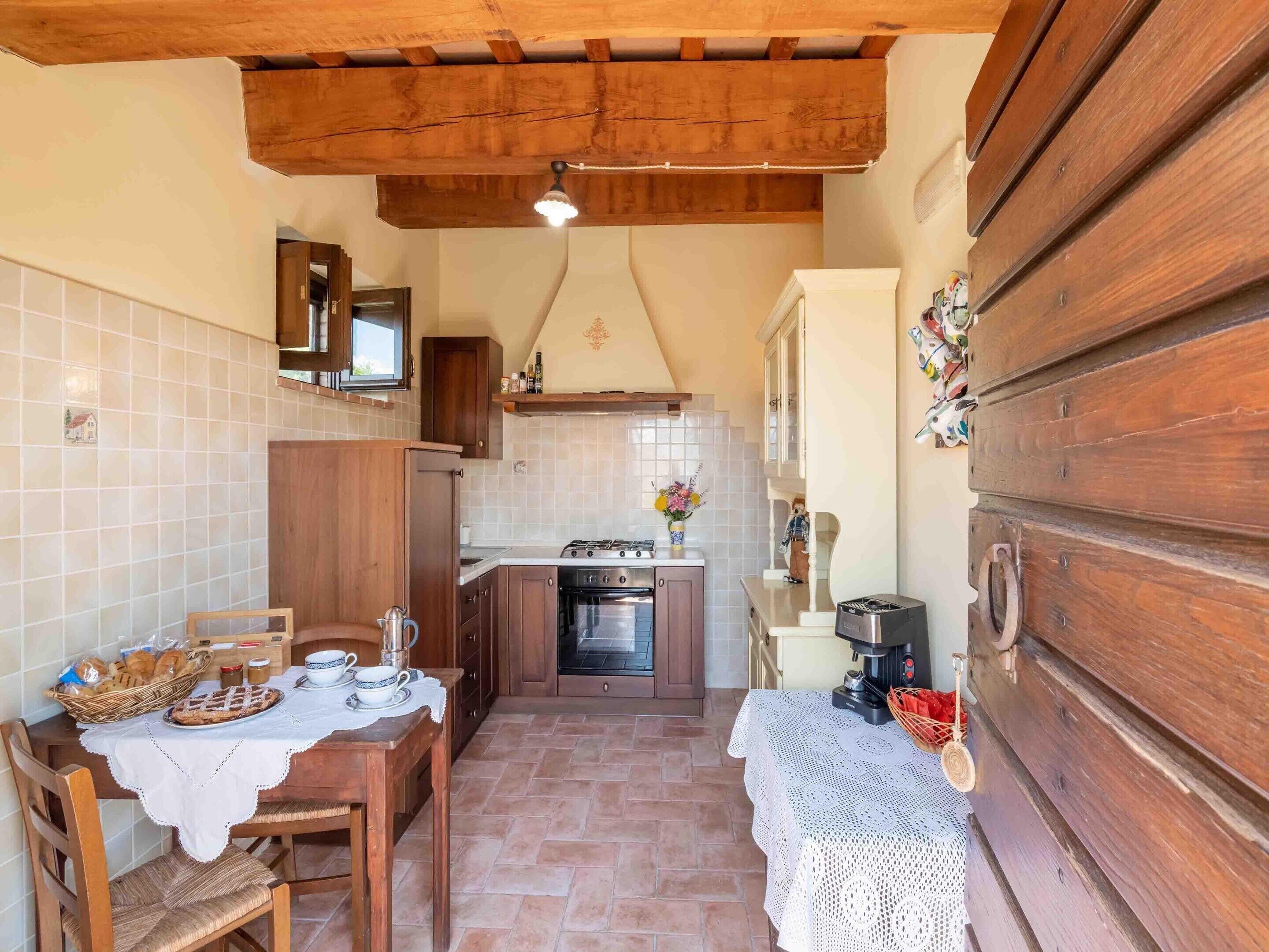 Una cocina compacta con armarios de madera, salpicadero azulejado, una mesa de comedor con mantel de encaje y un banco de madera.