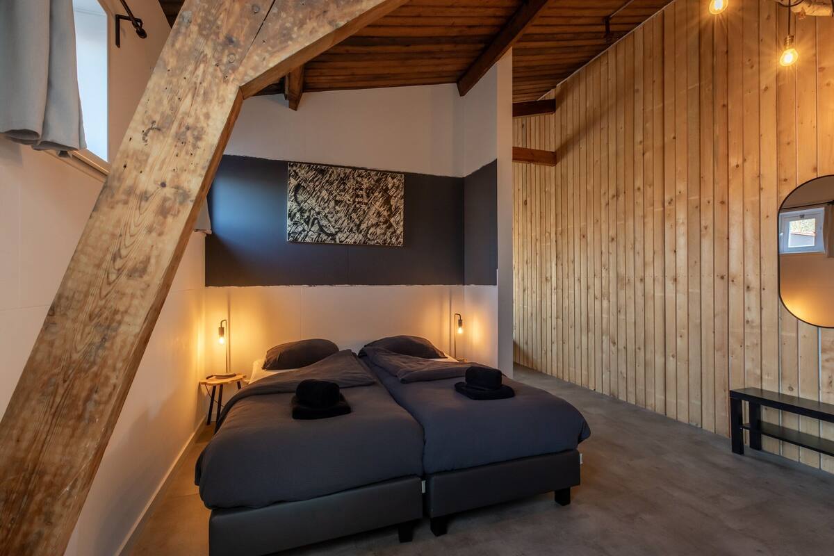 A loft-style bedroom with a sloped ceiling, two beds, a large abstract artwork, and a wooden wall.