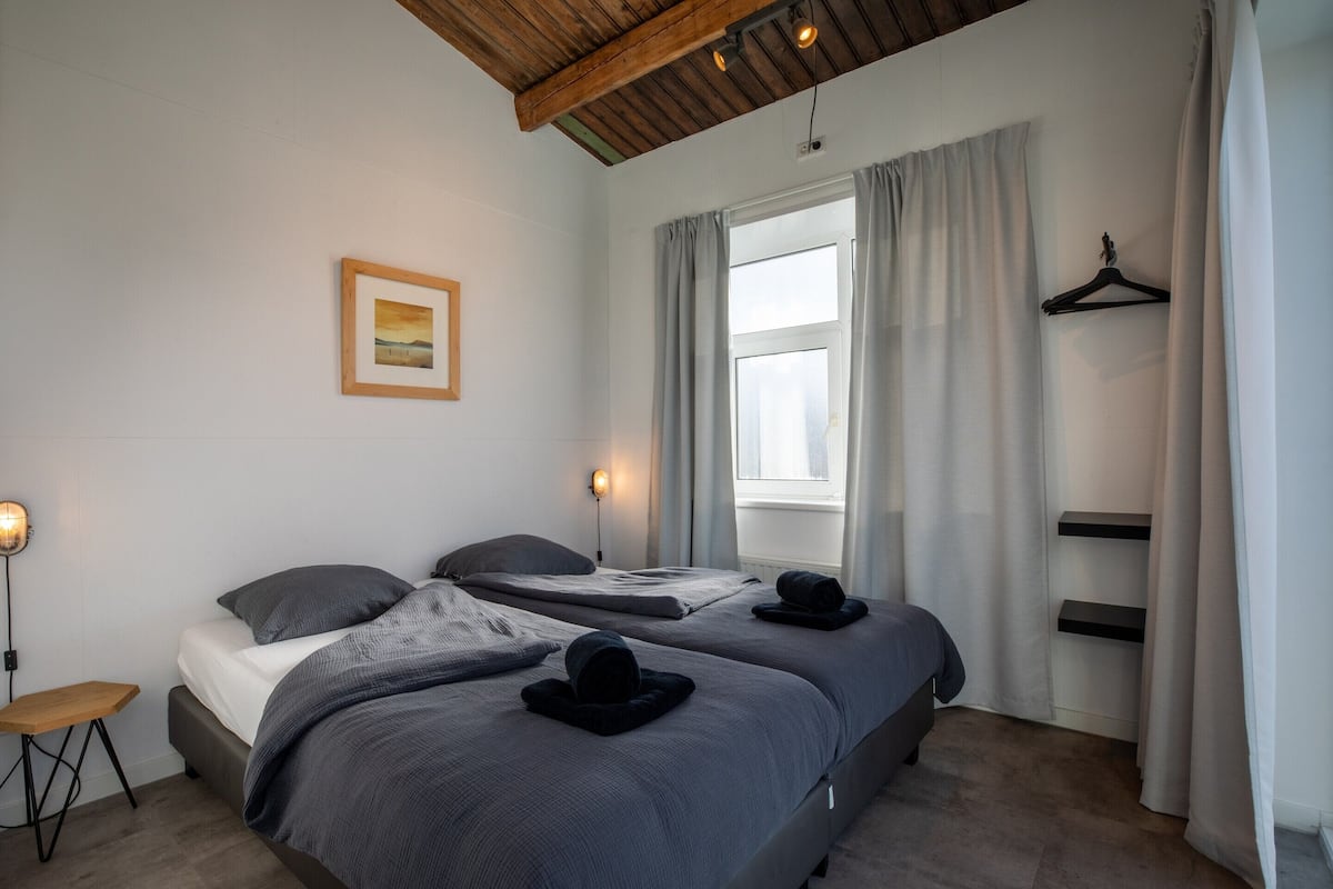 A bedroom with two beds, a wooden ceiling, a bedside table, a hanging light, a window with curtains, and a framed picture on the wall.